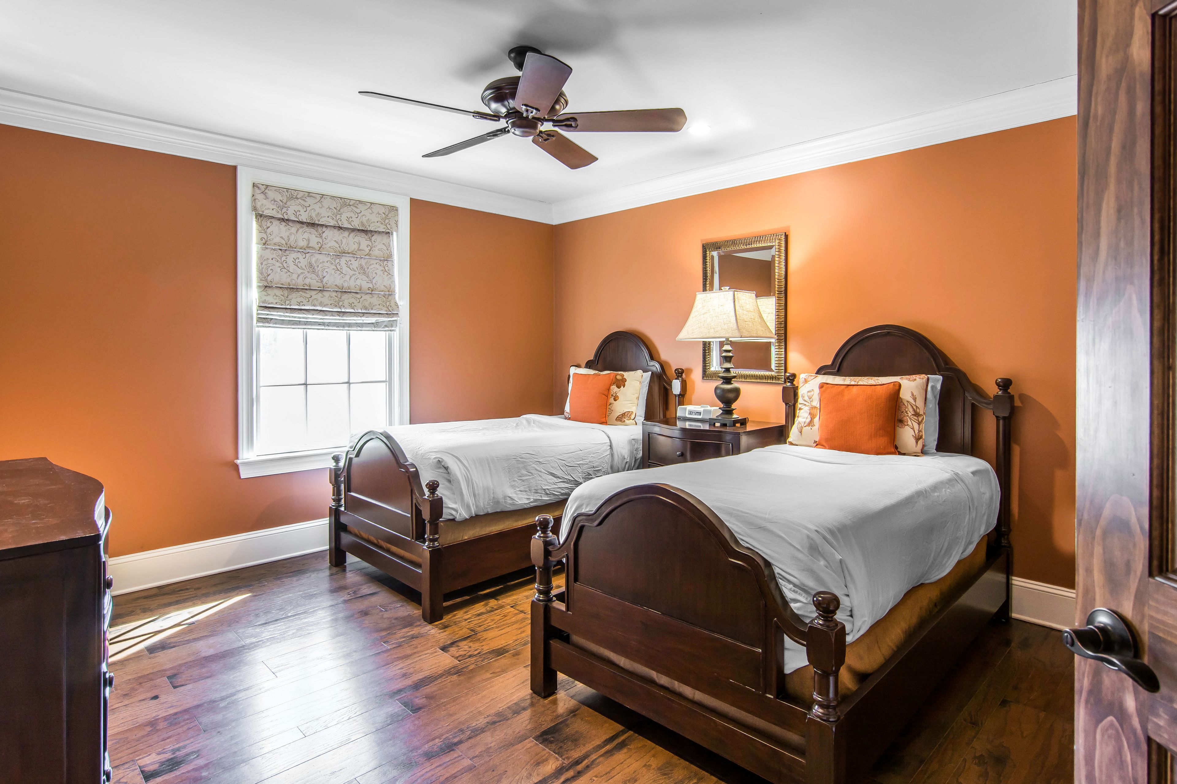 A bedroom with two wooden beds, a ceiling fan, a window with blinds, and a nightstand with a lamp.