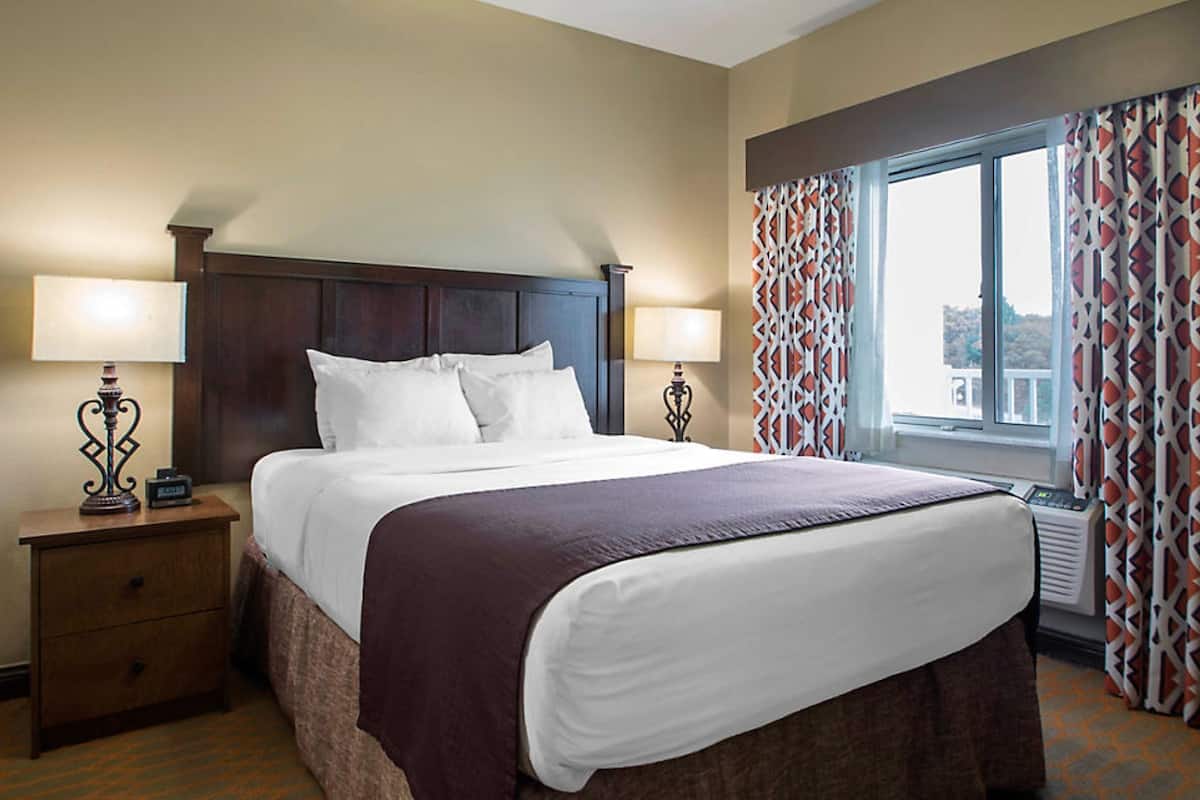 A hotel room with a large bed, two bedside tables with lamps, a window with patterned curtains, and a view of the city.