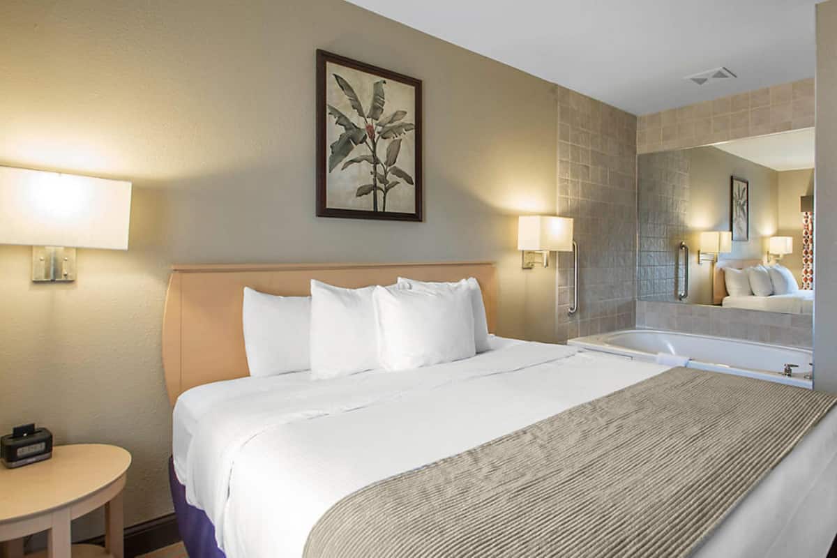 A hotel room with a large bed, a round table, a lamp, and a bathroom with a mirror and sink.