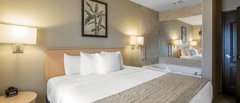 Habitación de hotel con una cama grande, una mesa redonda, una lámpara y un baño con espejo y lavamanos.