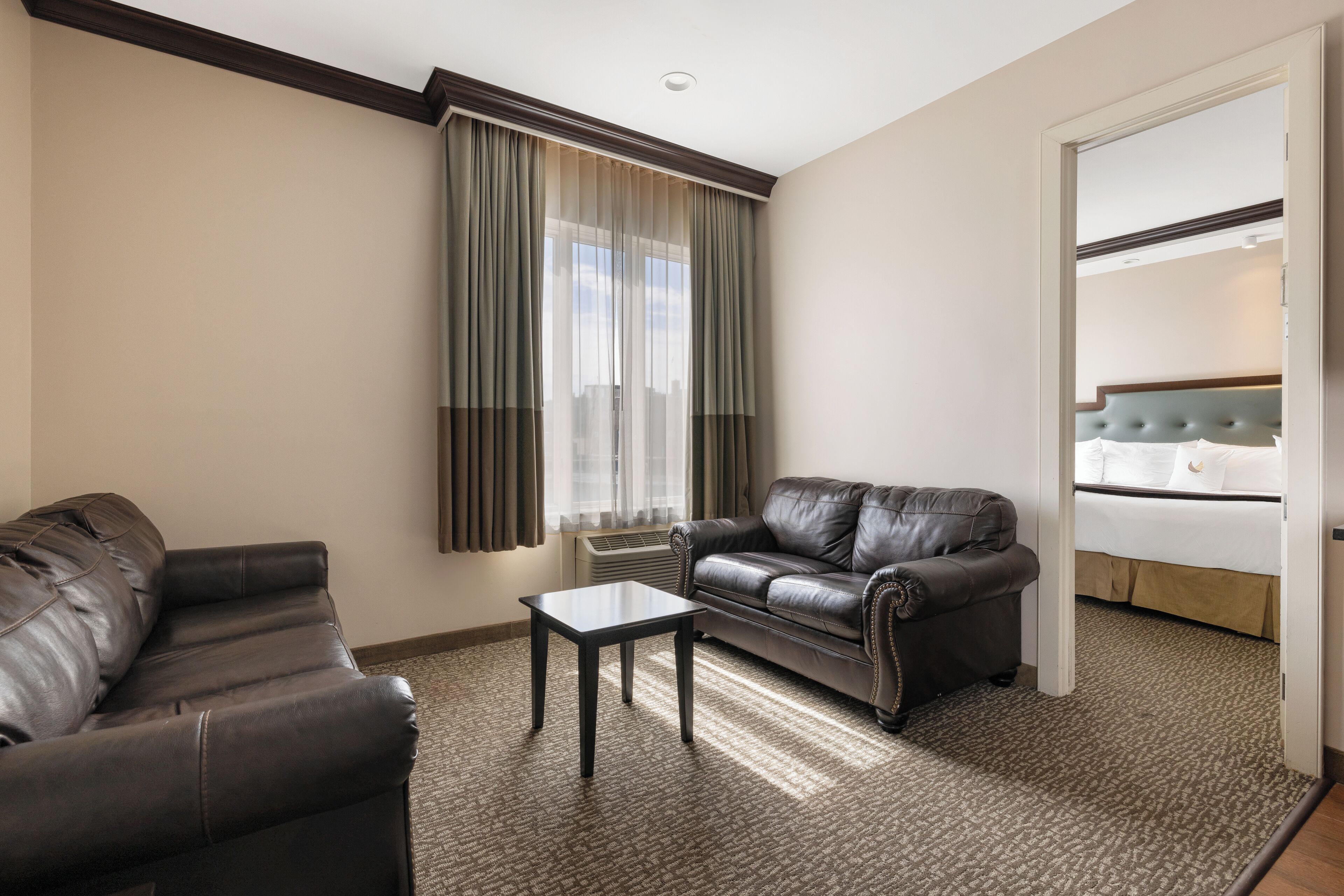 Una habitación de hotel con un sofá de cuero oscuro, una mesita y una cama con cabecera capitonada.