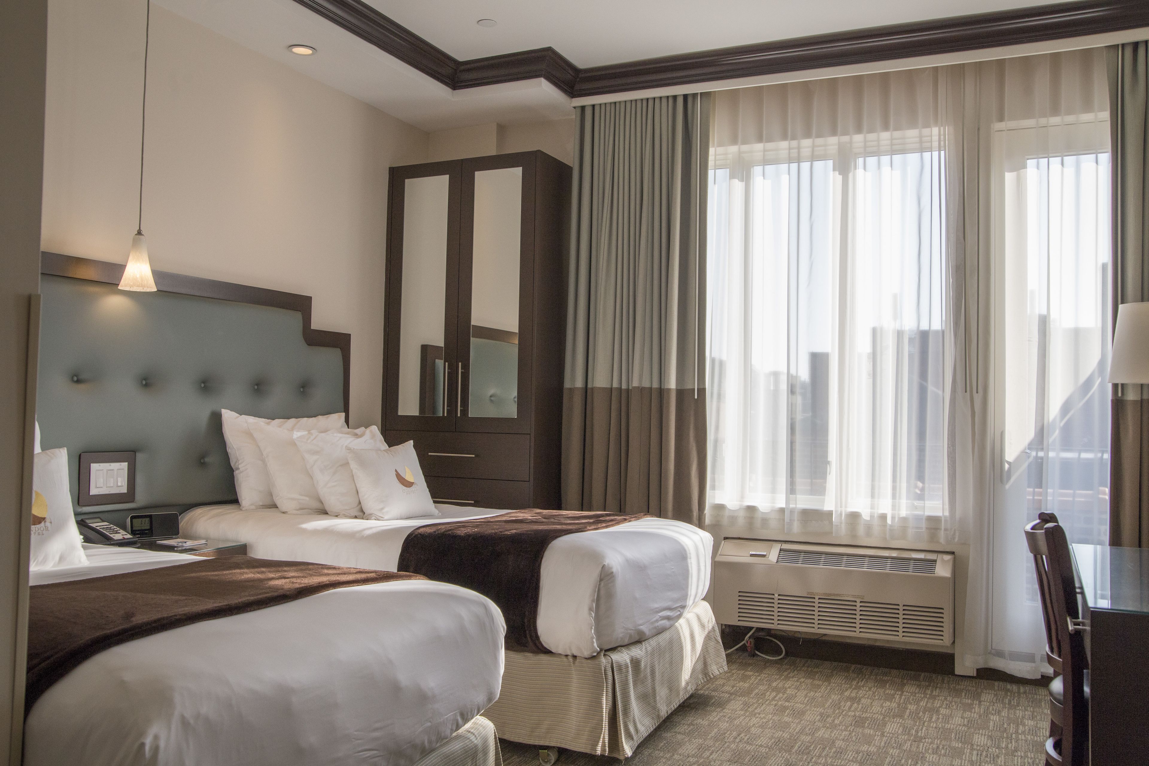 A hotel room with two beds, a large mirror, a desk, and a view of the city through large windows.