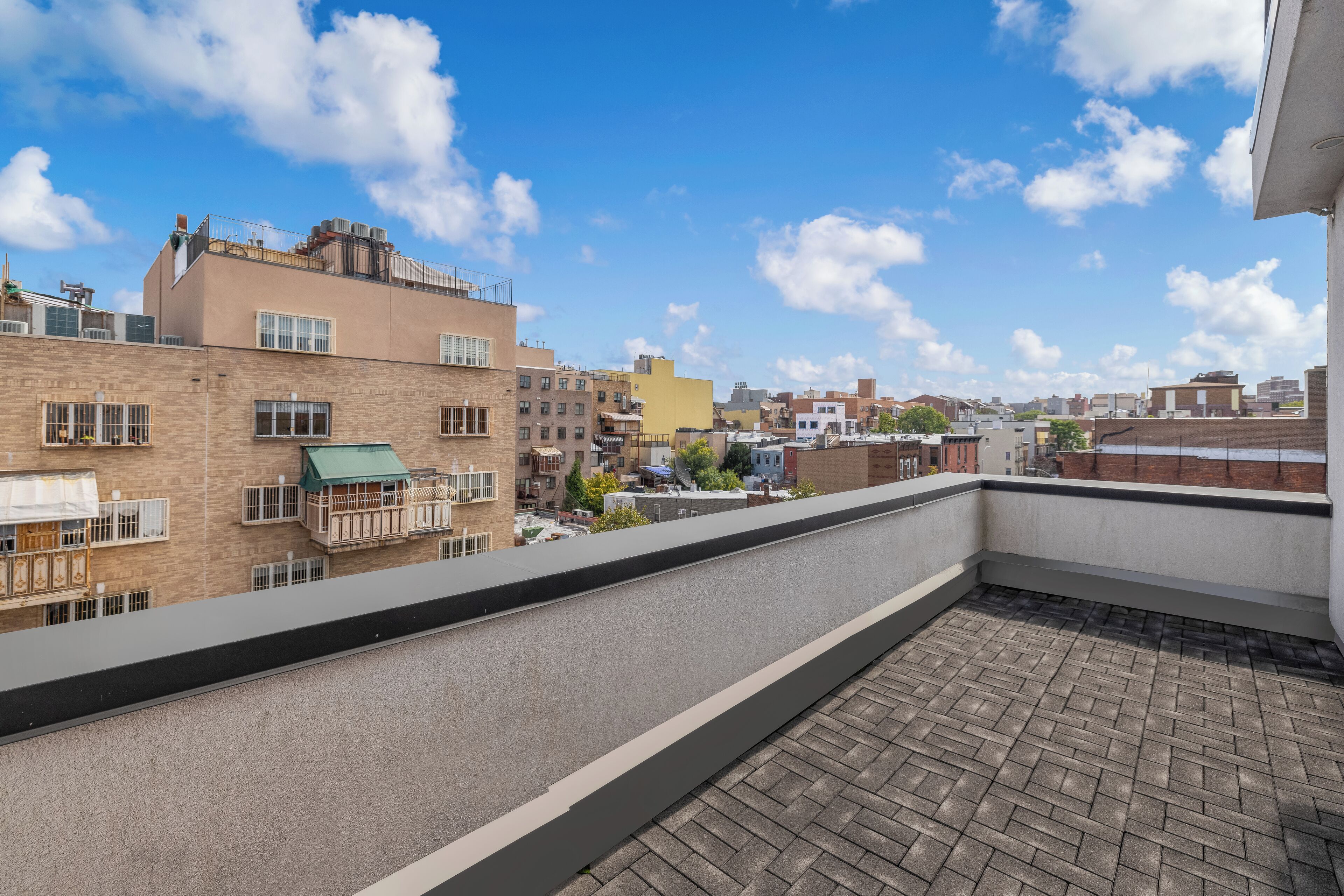 A rooftop terrace with a brick floor, overlooking a cityscape with various buildings and a clear blue sky.