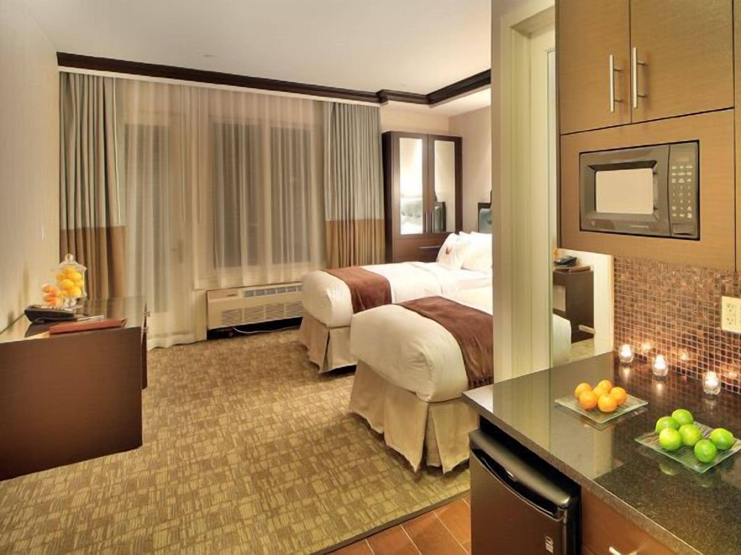 A hotel room with two beds, a desk, a microwave, and a small kitchen area with fruit on the counter.