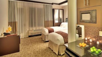 A hotel room with two beds, a desk, a microwave, and a small kitchen area with fruit on the counter.