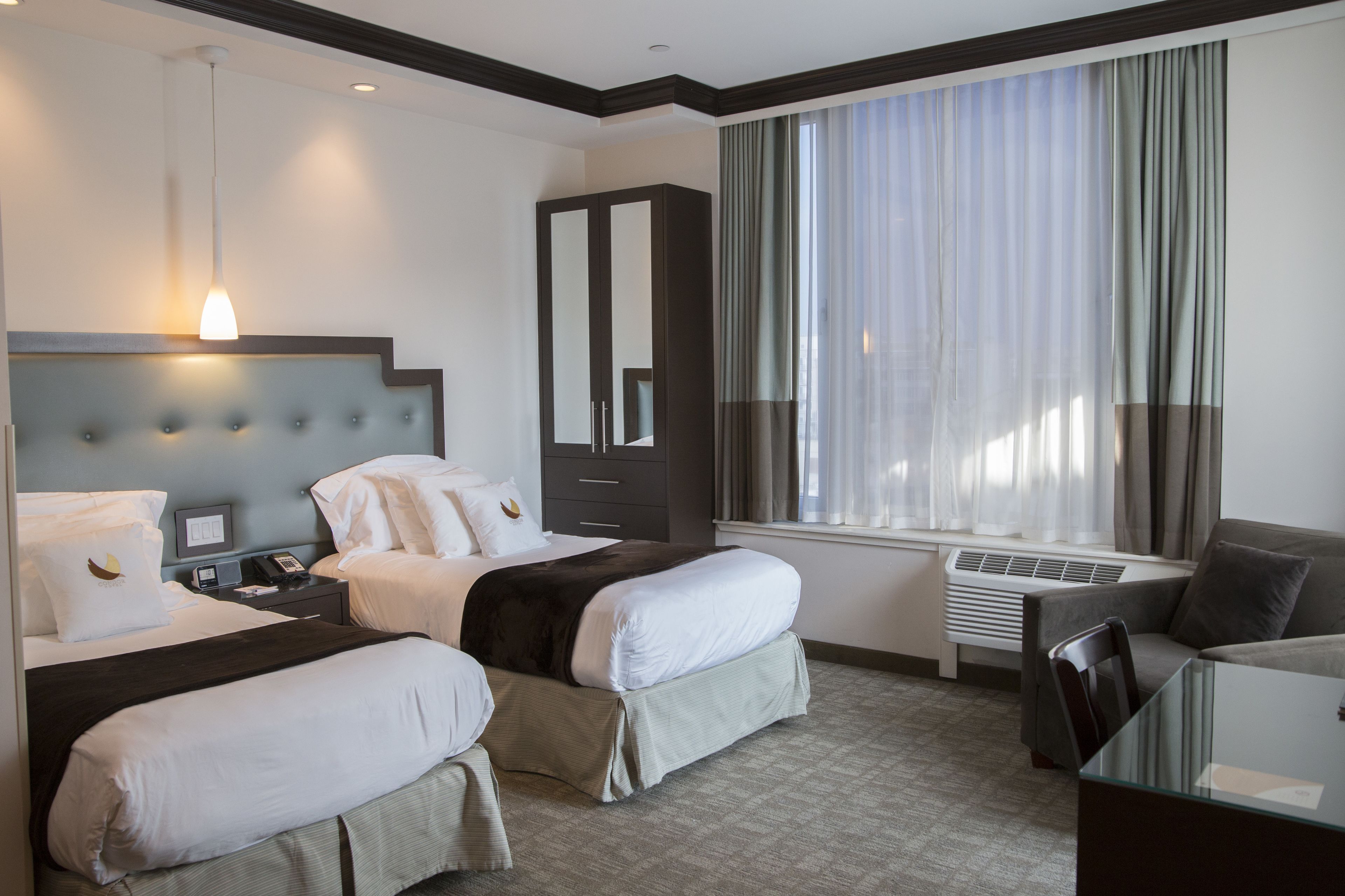 Quarto de hotel com duas camas, uma escrivaninha, uma cadeira, um guarda-roupa, uma janela grande com cortinas e um ar condicionado.