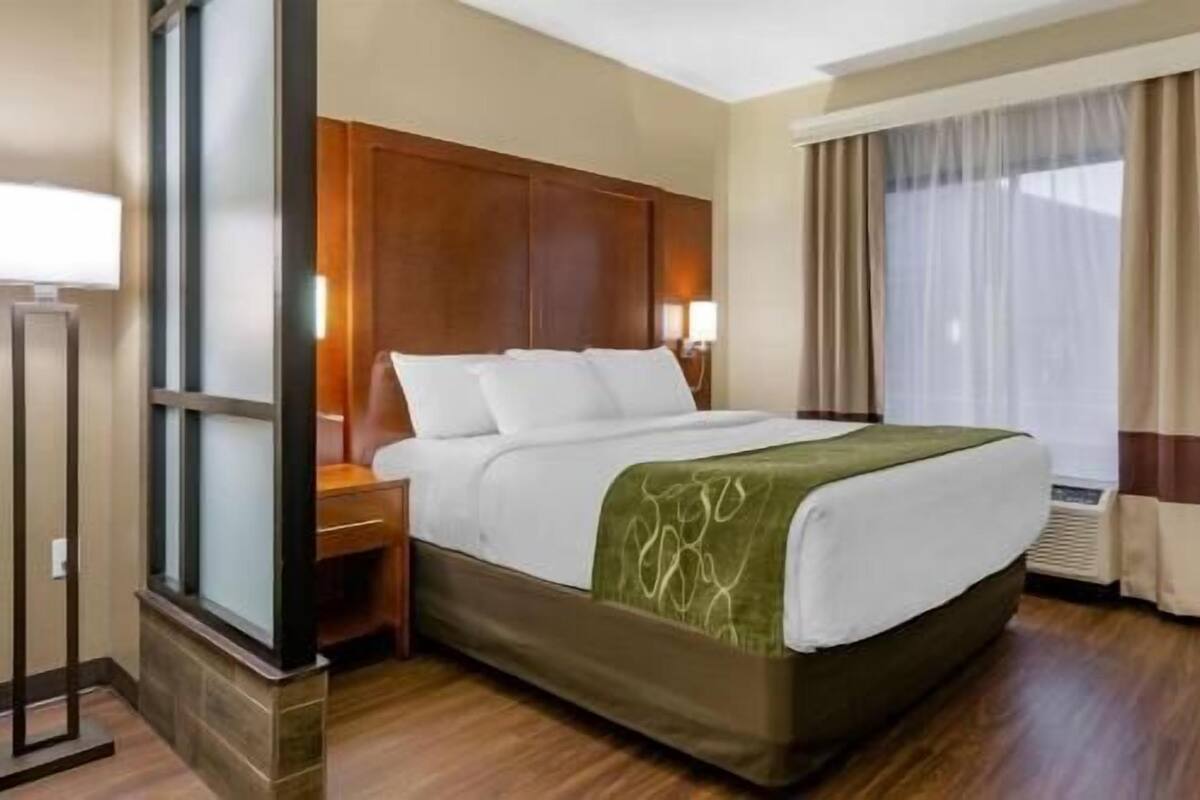 Ein Hotelzimmer mit einem Bett, einem Nachttisch, einer Lampe, einem Fenster mit Vorhängen und einem Wandheizkörper.