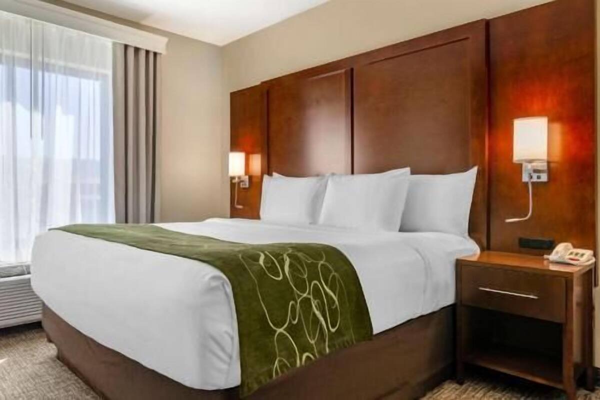Ein Hotelzimmer mit einem großen Bett, einem Nachttisch mit Lampe und einem Fenster mit Vorhängen.