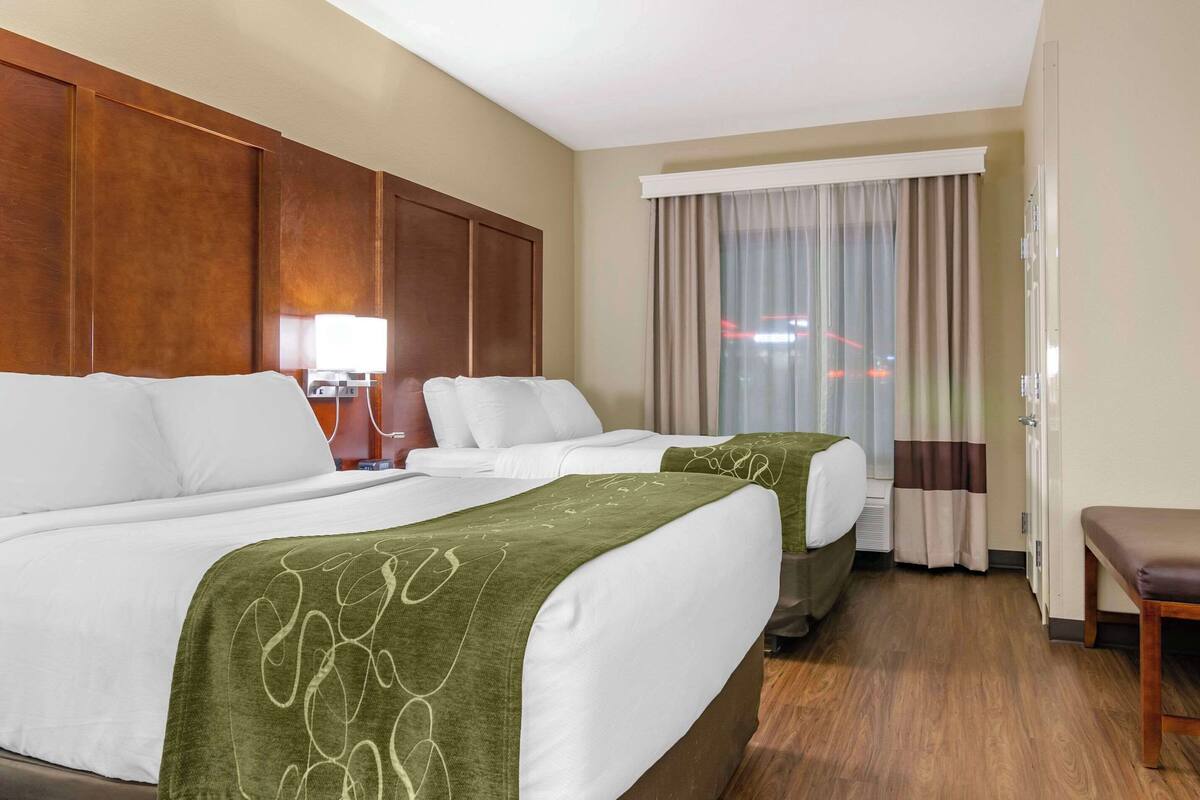 Ein Hotelzimmer mit zwei Betten, einem Holzkopfstück, einem Schreibtisch mit Lampe und Blick durch ein mit Vorhängen versehenes Fenster nach draußen.
