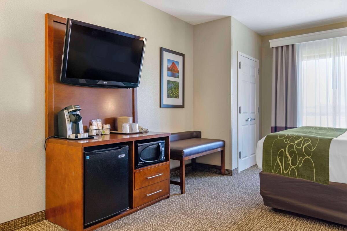 Ein Hotelzimmer mit Flachbildfernseher an der Wand, einer kleinen Kühlschrank, einer Mikrowelle, einem Schreibtisch mit Schubladen, einer Bank und einem Bett mit grüner Decke.