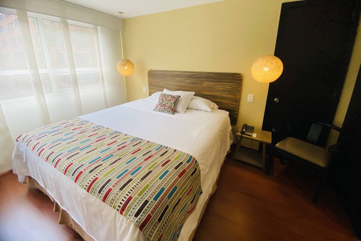 Una habitación de hotel con una cama grande, un escritorio con silla y un ventanal con cortinas.