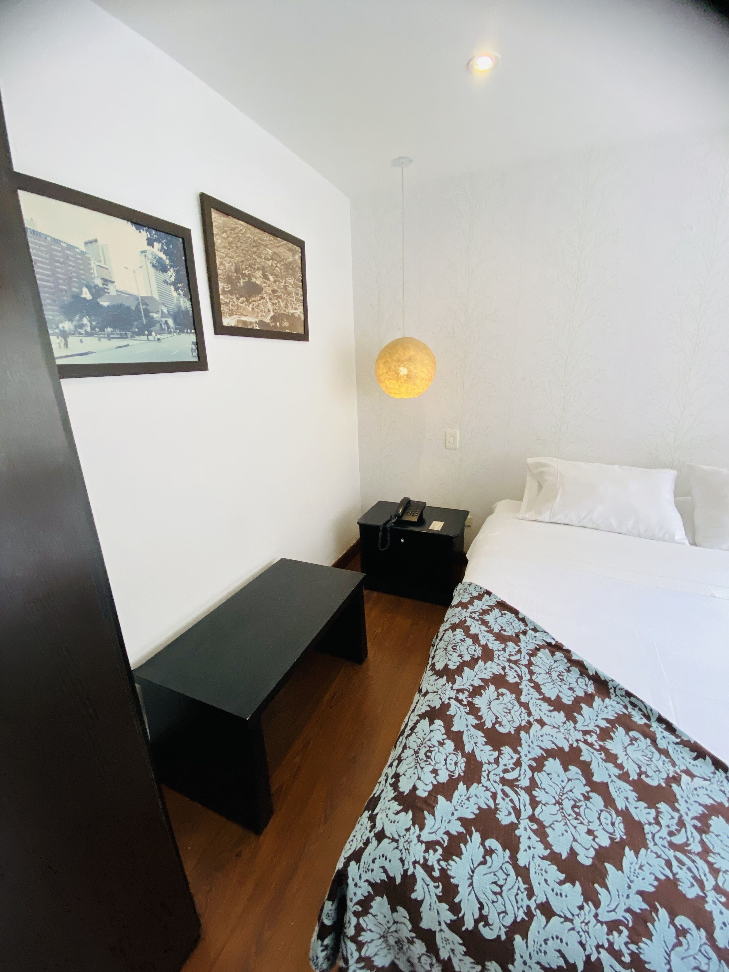 Quarto de hotel com cama, mesa de cabeceira, abajur e duas fotografias emolduradas na parede.
