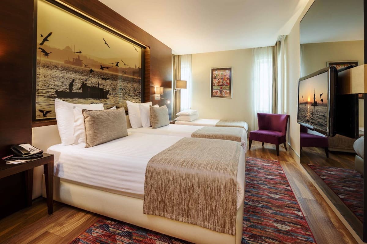 Une chambre d’hôtel avec un grand lit, un lit plus petit, un bureau avec un téléphone, une chaise, une télévision et un tableau représentant des oiseaux et un bateau.