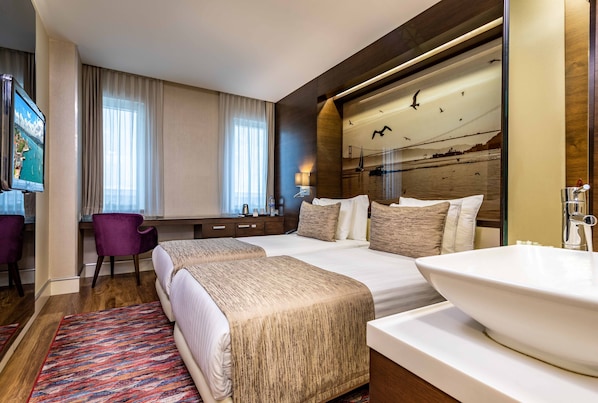 A hotel room with two beds, a large window with curtains, a desk with a chair, a television, and a large sink.