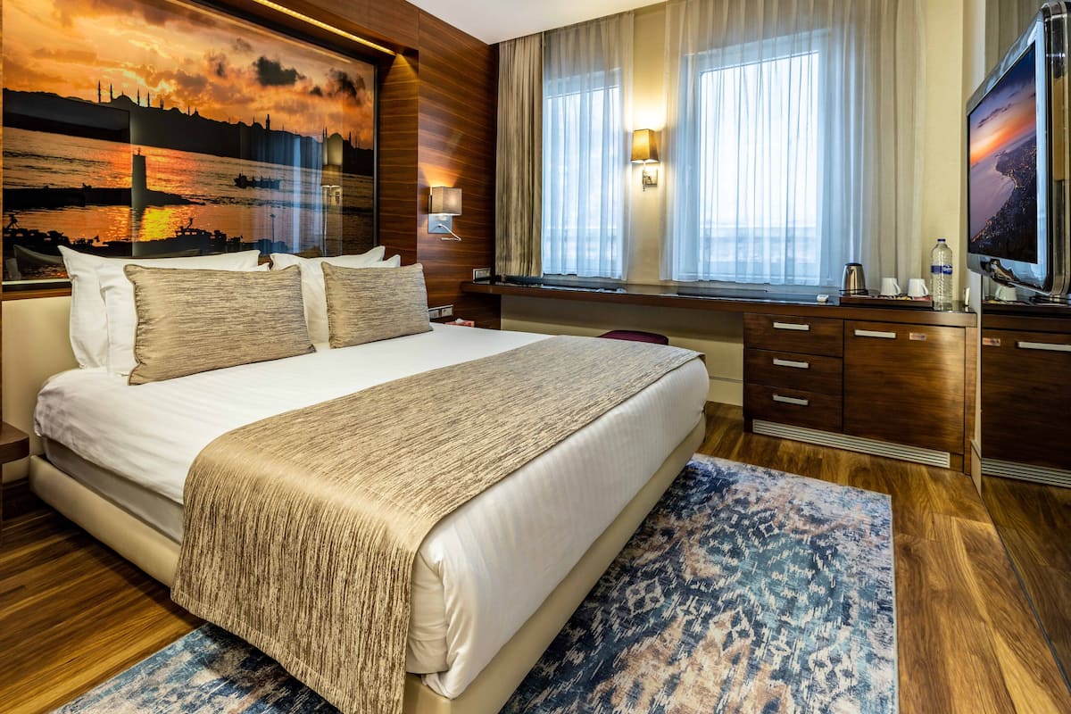 Une chambre d’hôtel avec un grand lit, un bureau avec un ordinateur, une télévision et un tableau représentant un coucher de soleil sur l’horizon d’une ville.