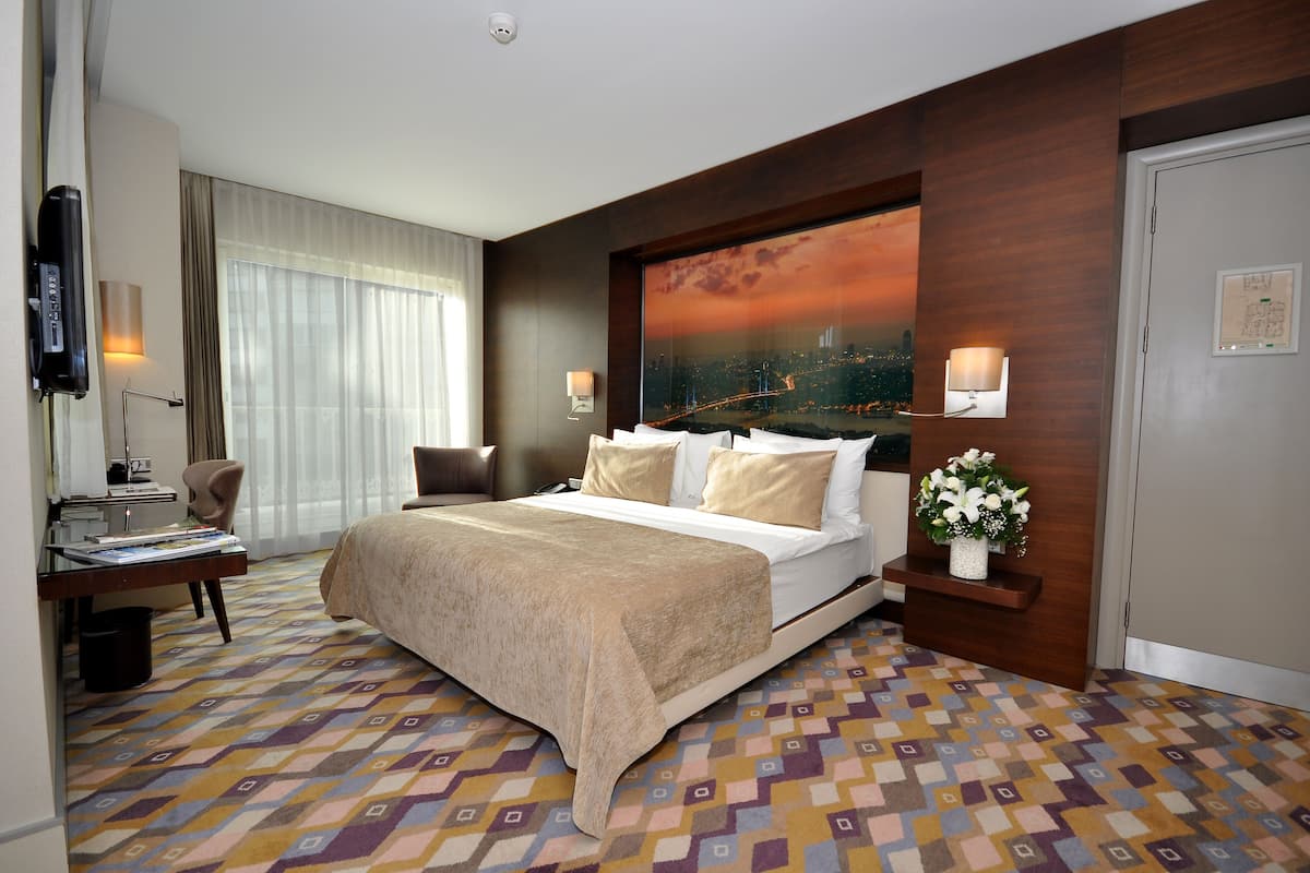Une chambre d’hôtel avec un lit, un bureau, une chaise, une lampe et une vue sur le paysage urbain par la fenêtre.