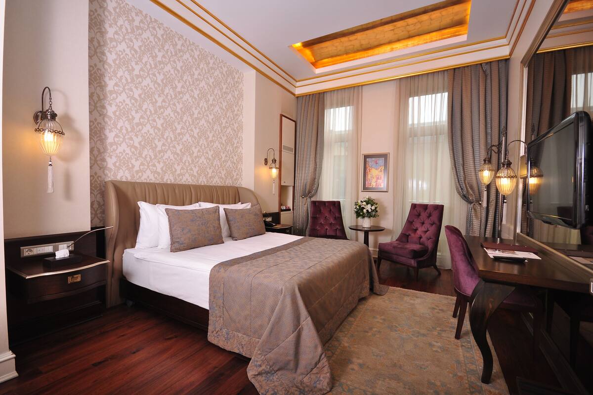 Une chambre d’hôtel avec un grand lit, un bureau avec une chaise, une télévision et une petite table sur laquelle est posé un vase de fleurs.