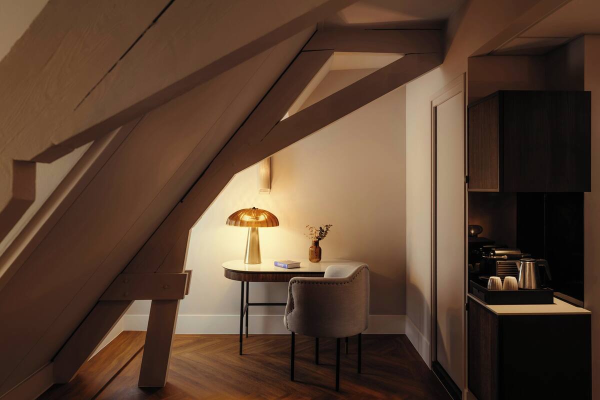 A cozy loft bedroom with a sloped ceiling, a small round table with a lamp, a chair, and a coffee machine.