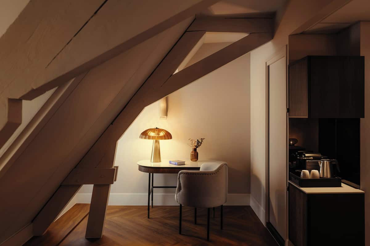 A cozy loft bedroom with a sloped ceiling, a small round table with a lamp, a chair, and a coffee machine.