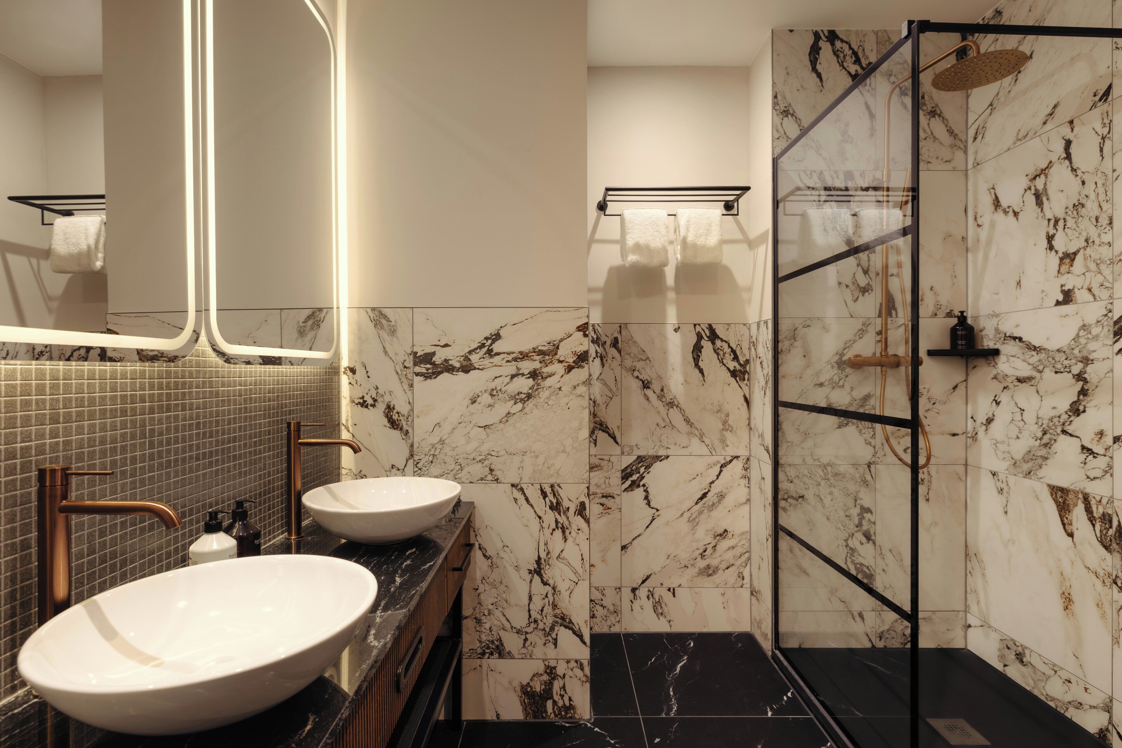 Un baño moderno con dos lavamanos, ducha con mampara de vidrio y paredes de mármol.