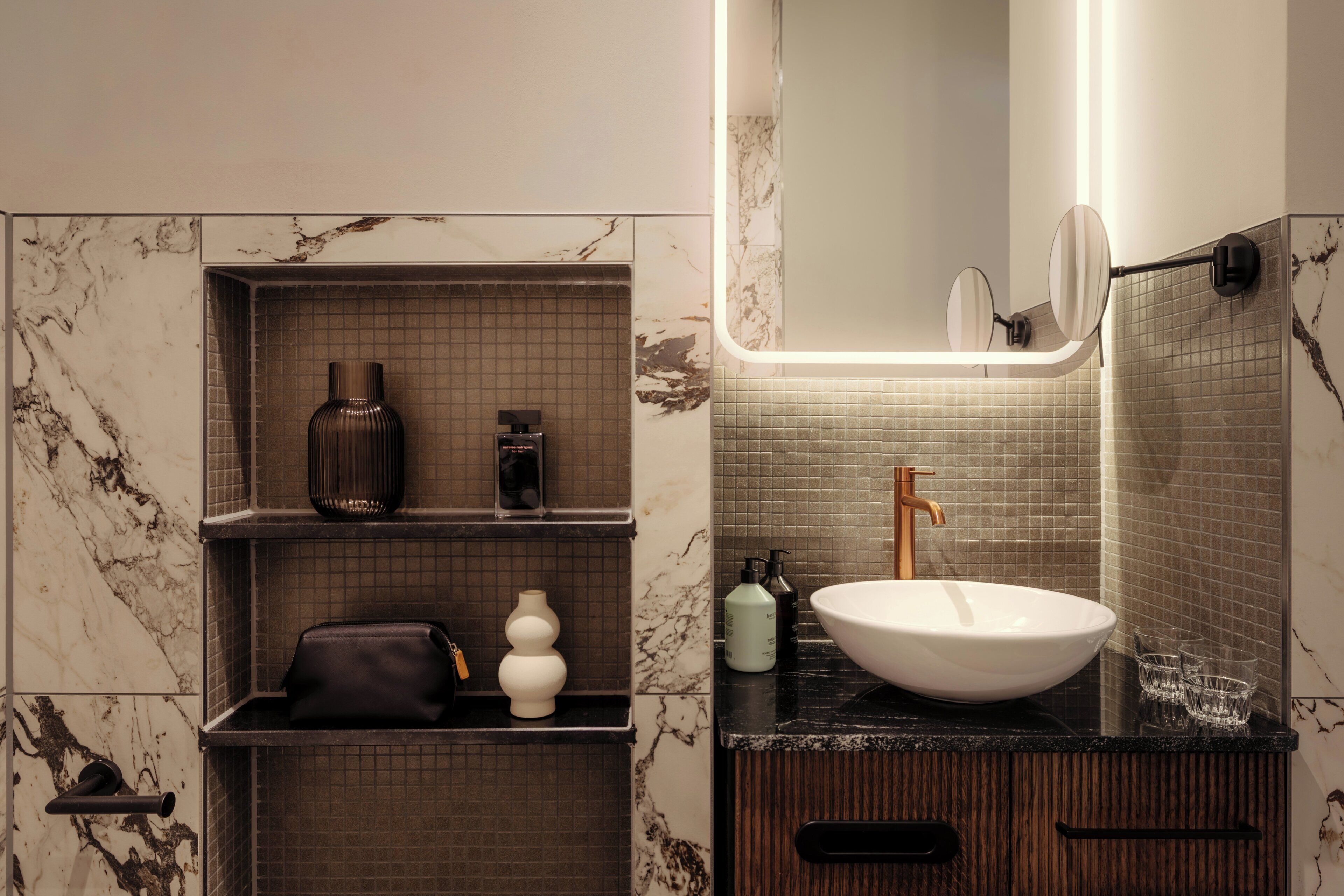 Un baño con pared de mármol, encimera negra, lavabo blanco y un espejo.
