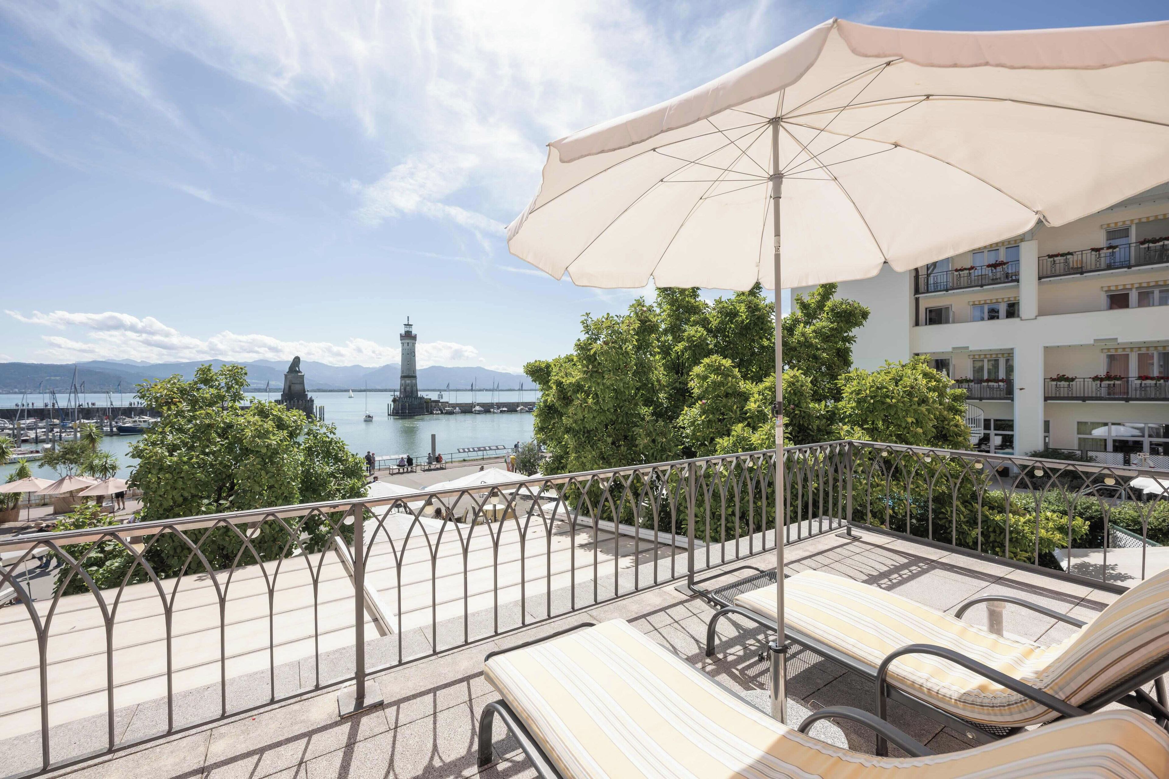 A balcony with lounge chairs and an umbrella overlooking a marina with boats and a lighthouse.