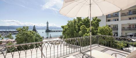 Un balcon doté de chaises longues et d’un parasol, offrant une vue sur une marina avec des bateaux et un phare.