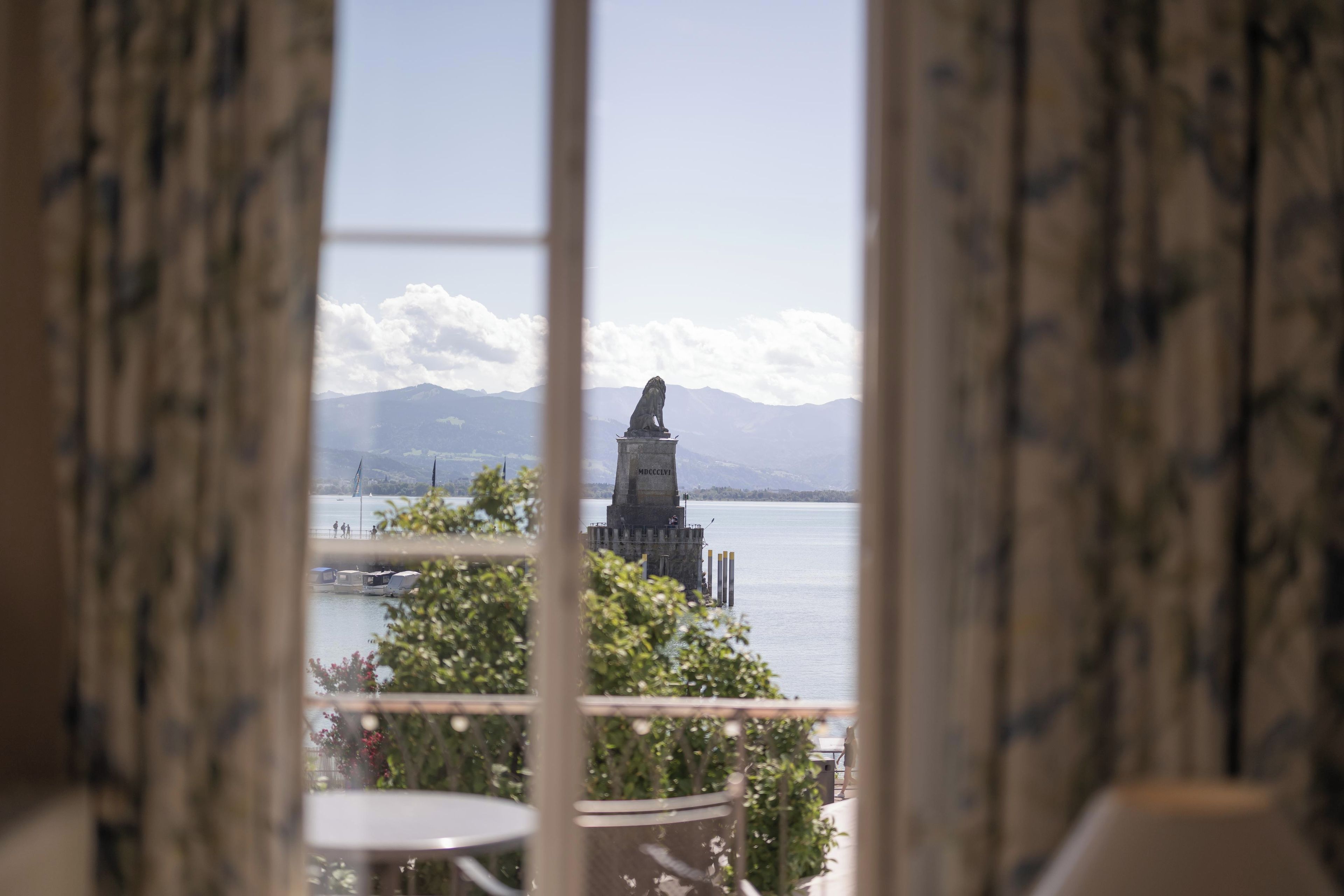 A view through a window with curtains, featuring a statue on a pedestal by a body of water, with mountains in the background.
