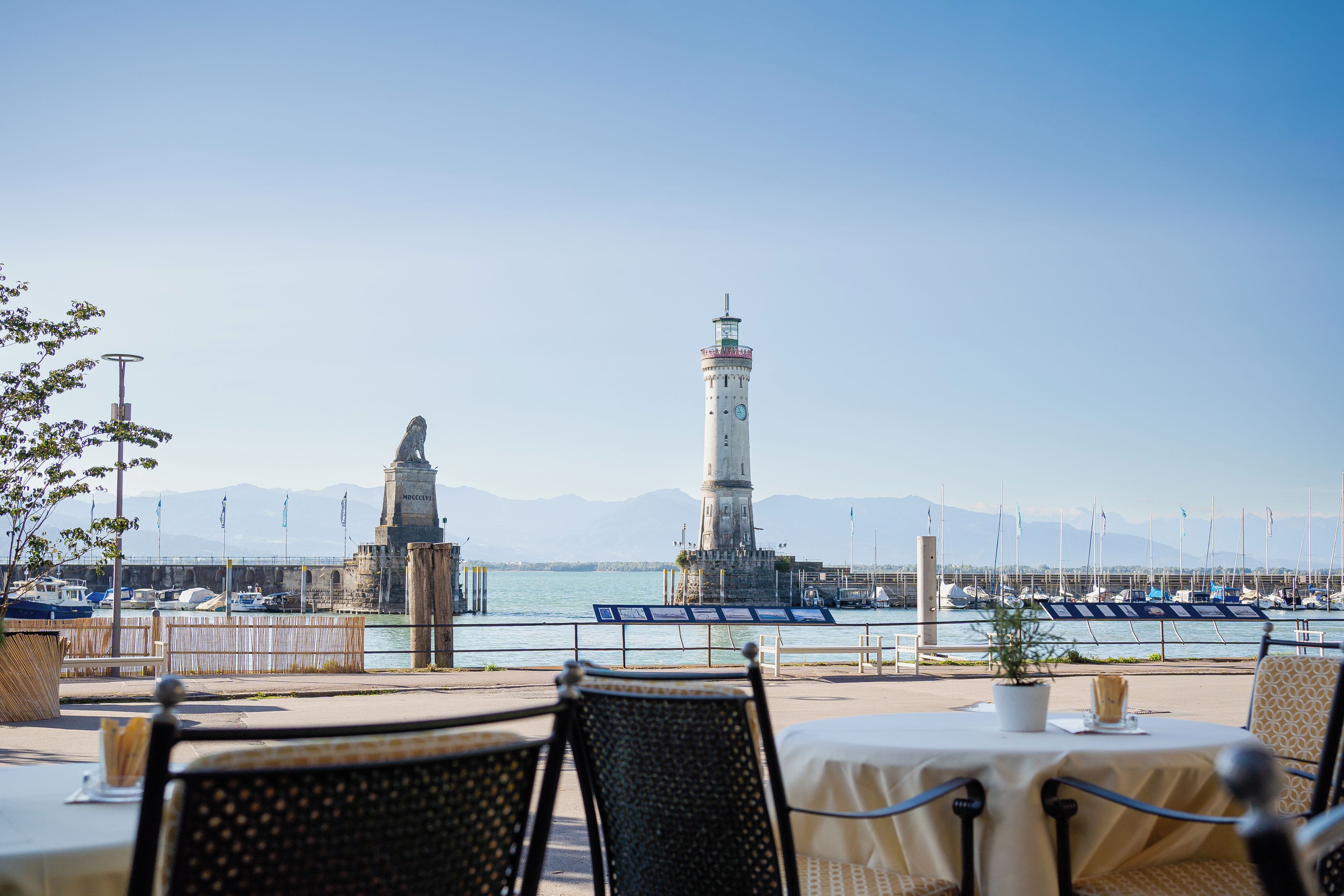 A waterfront dining area with tables and chairs, a statue, and a lighthouse in the background.