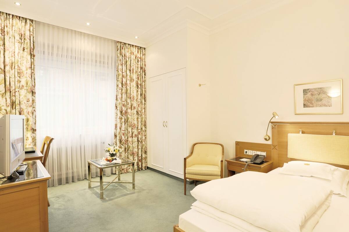 A hotel room with a bed, a desk with a computer, a chair, a nightstand with a phone, and a window with curtains.