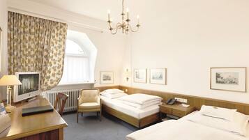 Quarto de hotel com cama, escrivaninha, cadeira e uma janela grande com cortinas.