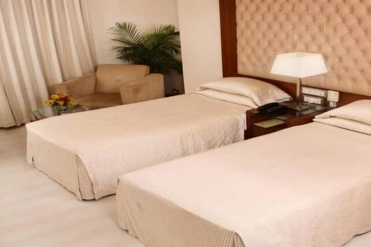 Quarto de hotel com duas camas de solteiro, uma mesa de cabeceira com abajur, uma cadeira e um vaso com planta.