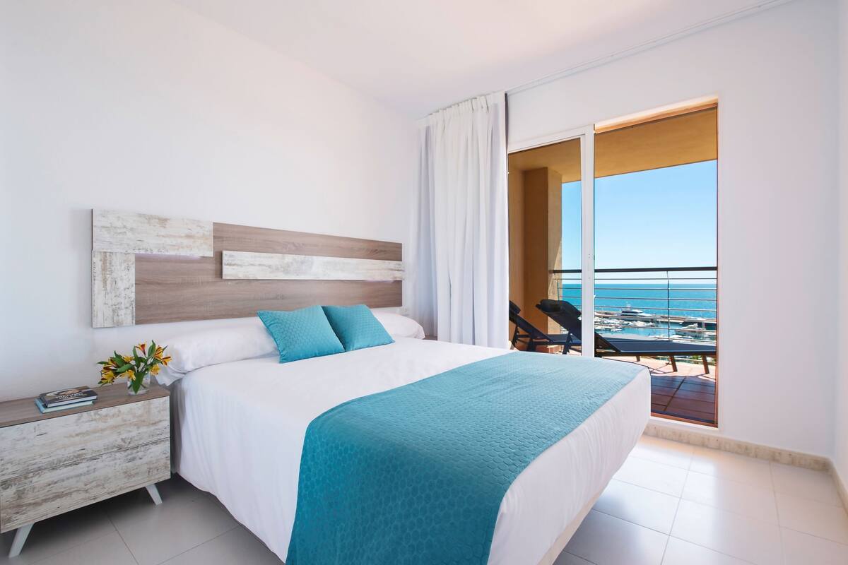 A bedroom with a large bed, a nightstand, and a view of the ocean through an open door.