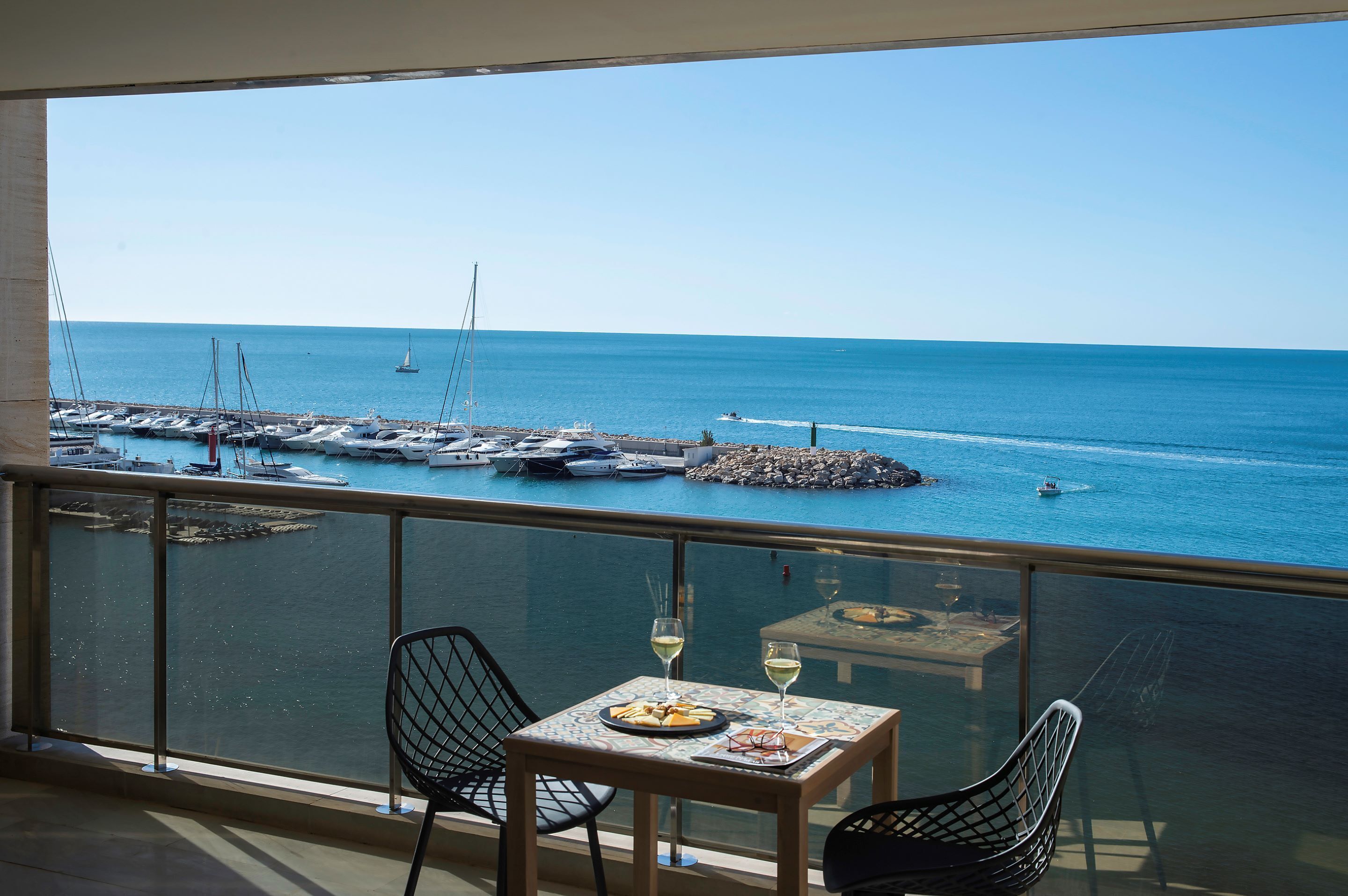A balcony with a table set for two, overlooking a marina with boats and a clear blue sea.
