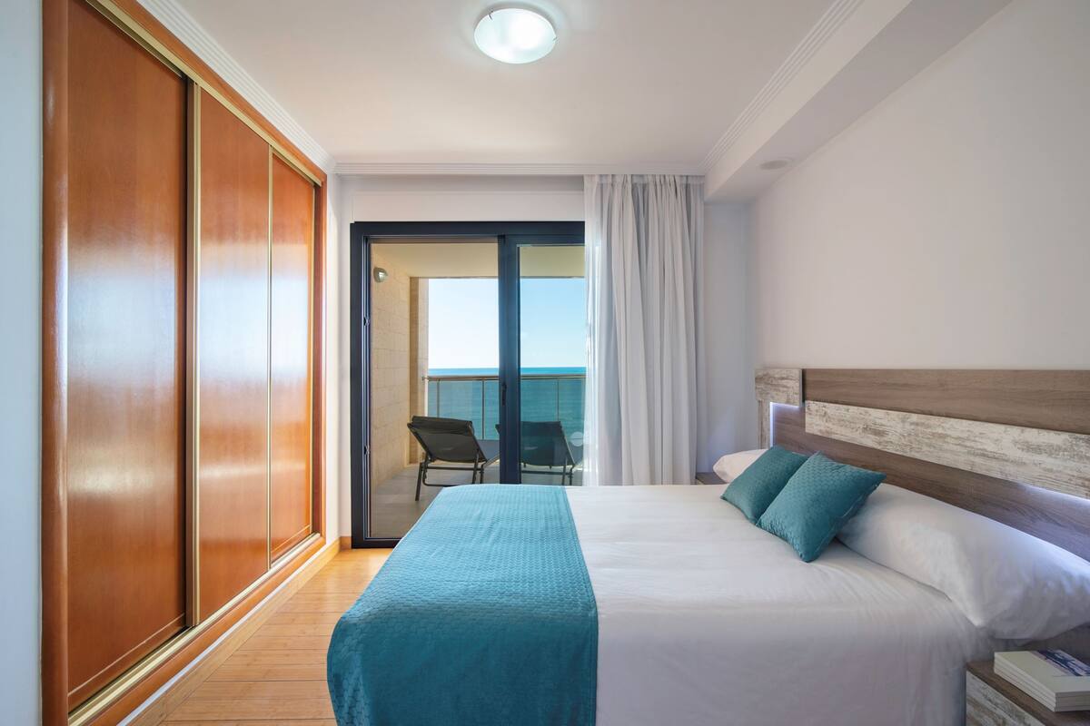 A hotel room with a bed, a wooden wardrobe, a balcony with a chair, and a view of the sea.