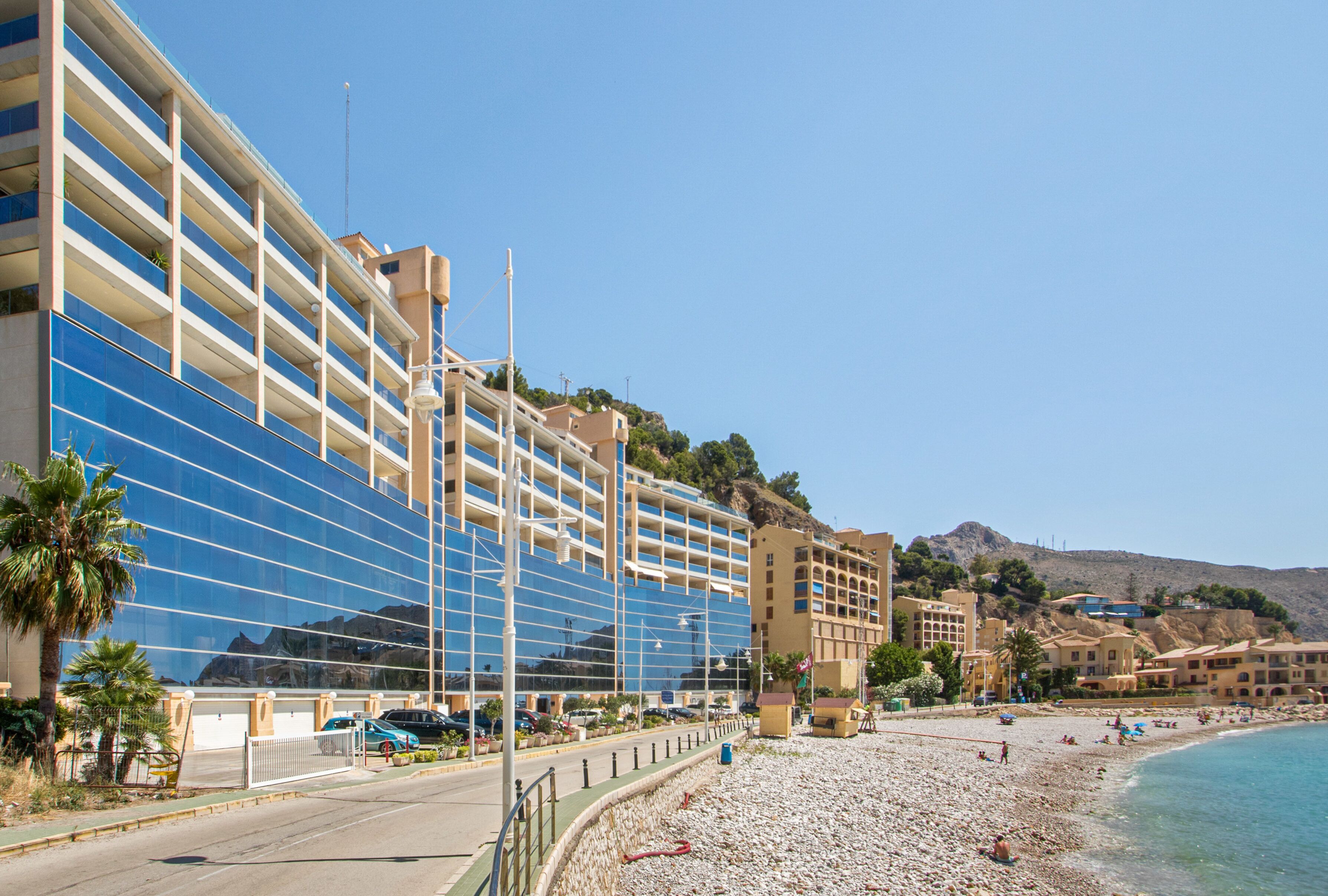 A modern hotel with a reflective facade, a beachfront location, and a rocky hillside in the background.