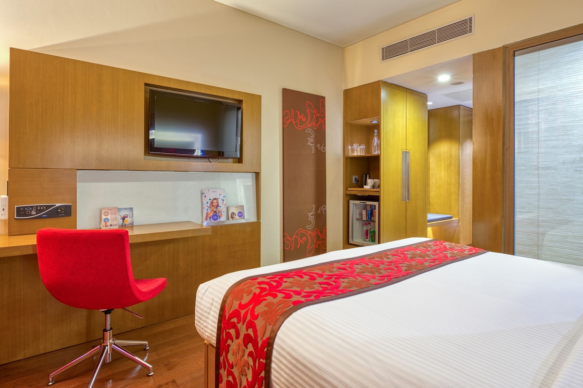A hotel room with a bed, a red chair, a TV, a shelf with toiletries, and a wooden wardrobe.