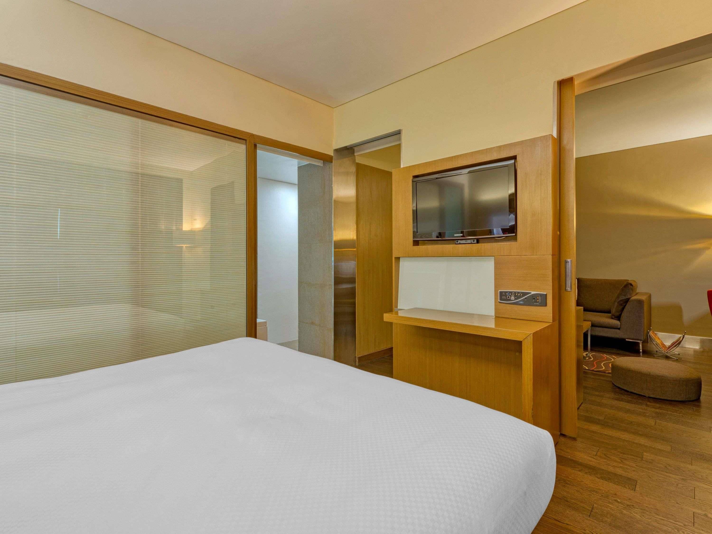 Una habitación de hotel moderna con una cama grande, un televisor de pantalla plana montado en una consola de madera y un vidrio corredizo que da acceso a un baño.