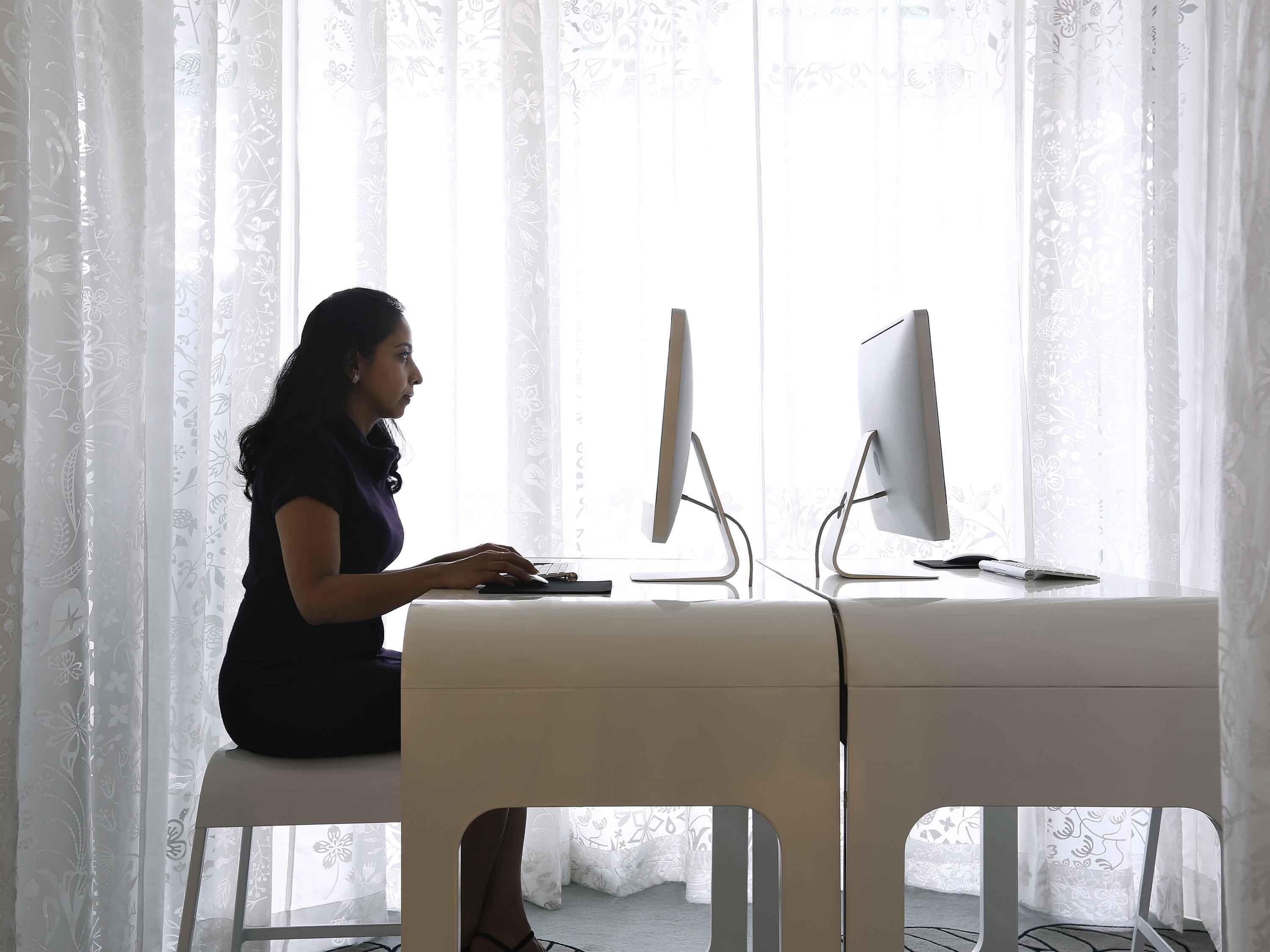 A person seated at a desk with two monitors, a keyboard, and a mouse. The room has a window with sheer curtains and a chair with a cut-out backrest.