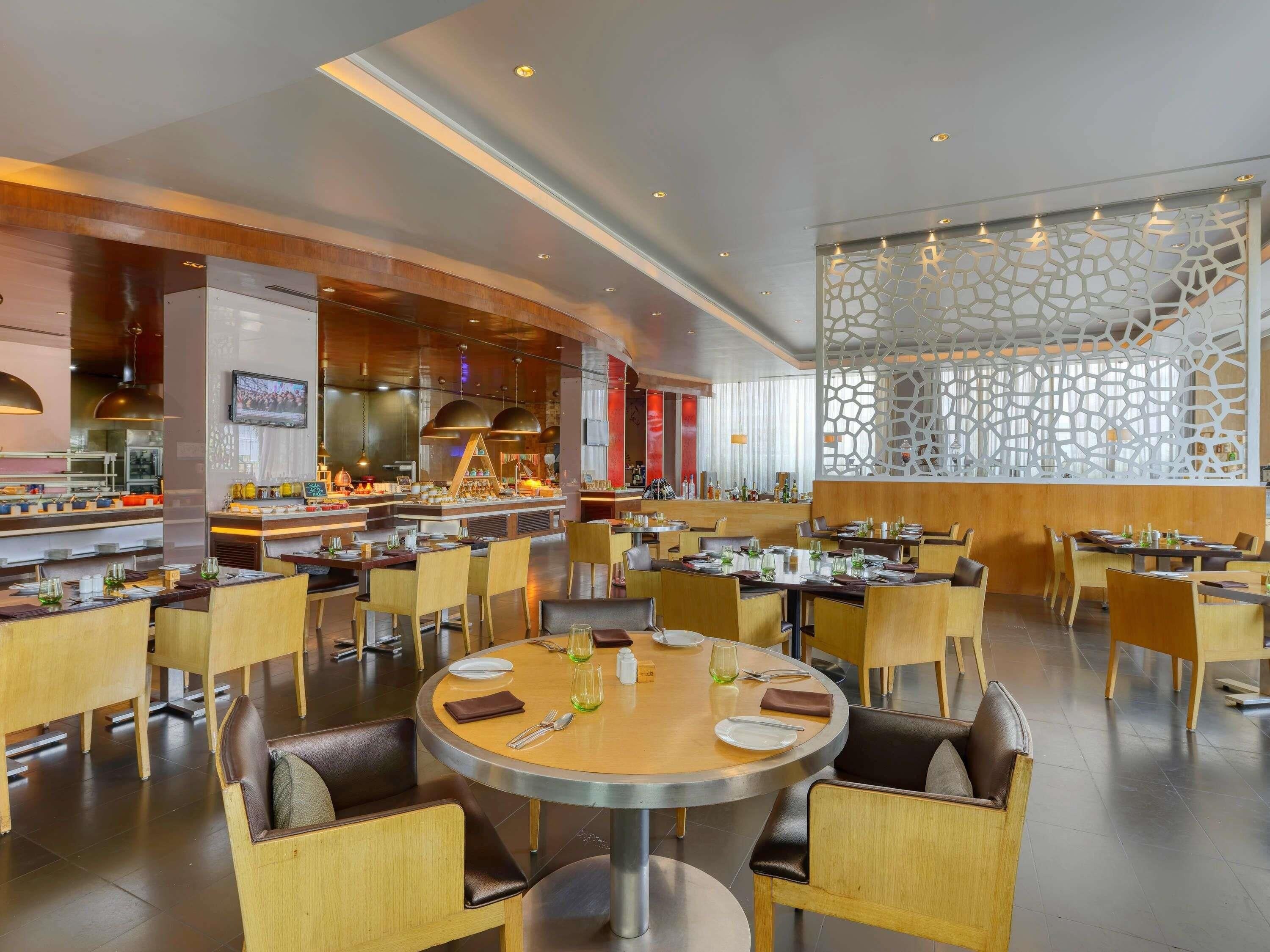 A modern restaurant with round tables, wooden chairs, and a kitchen in the background.