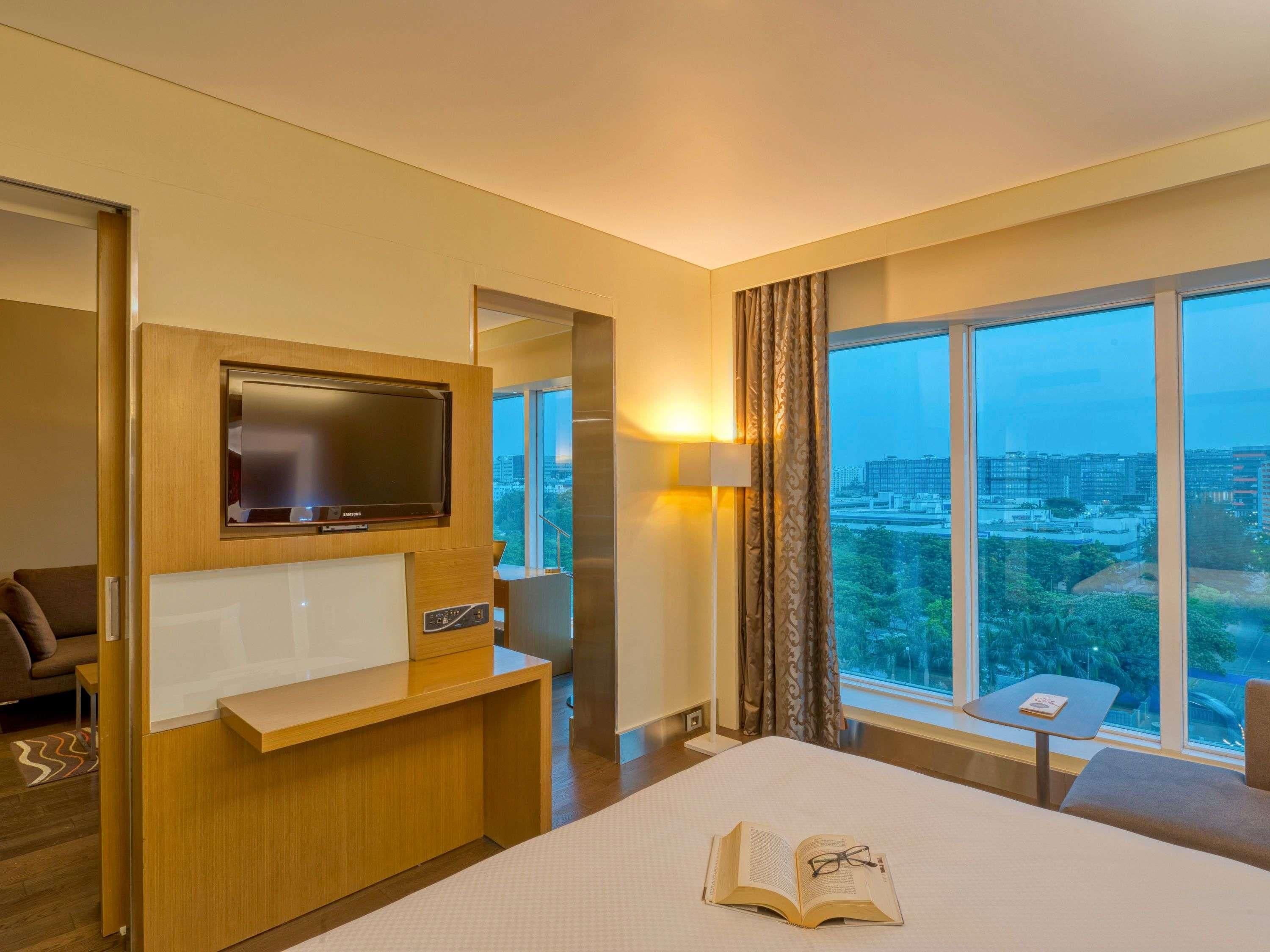 A hotel room with a bed, a TV, a desk, a chair, a small table, and a view of the city through large windows.