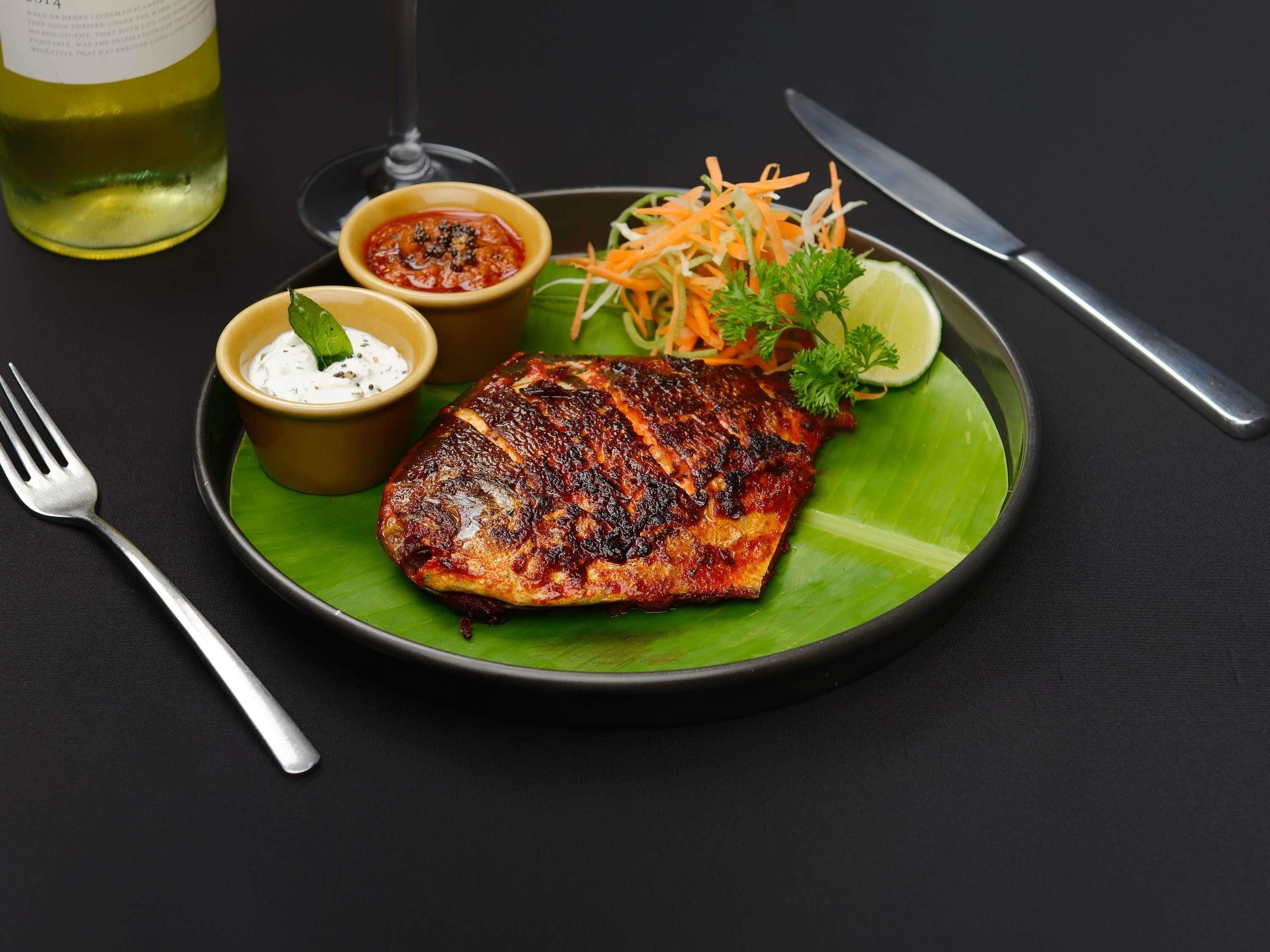 Ein Teller mit gegrilltem Fisch, einer Gabel, einem Messer und zwei kleinen Soßenbechern auf dunklem Untergrund.