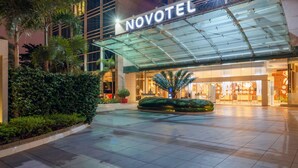 Entrada del hotel Novotel con un toldo de cristal, un vestíbulo bien iluminado y una zona ajardinada.