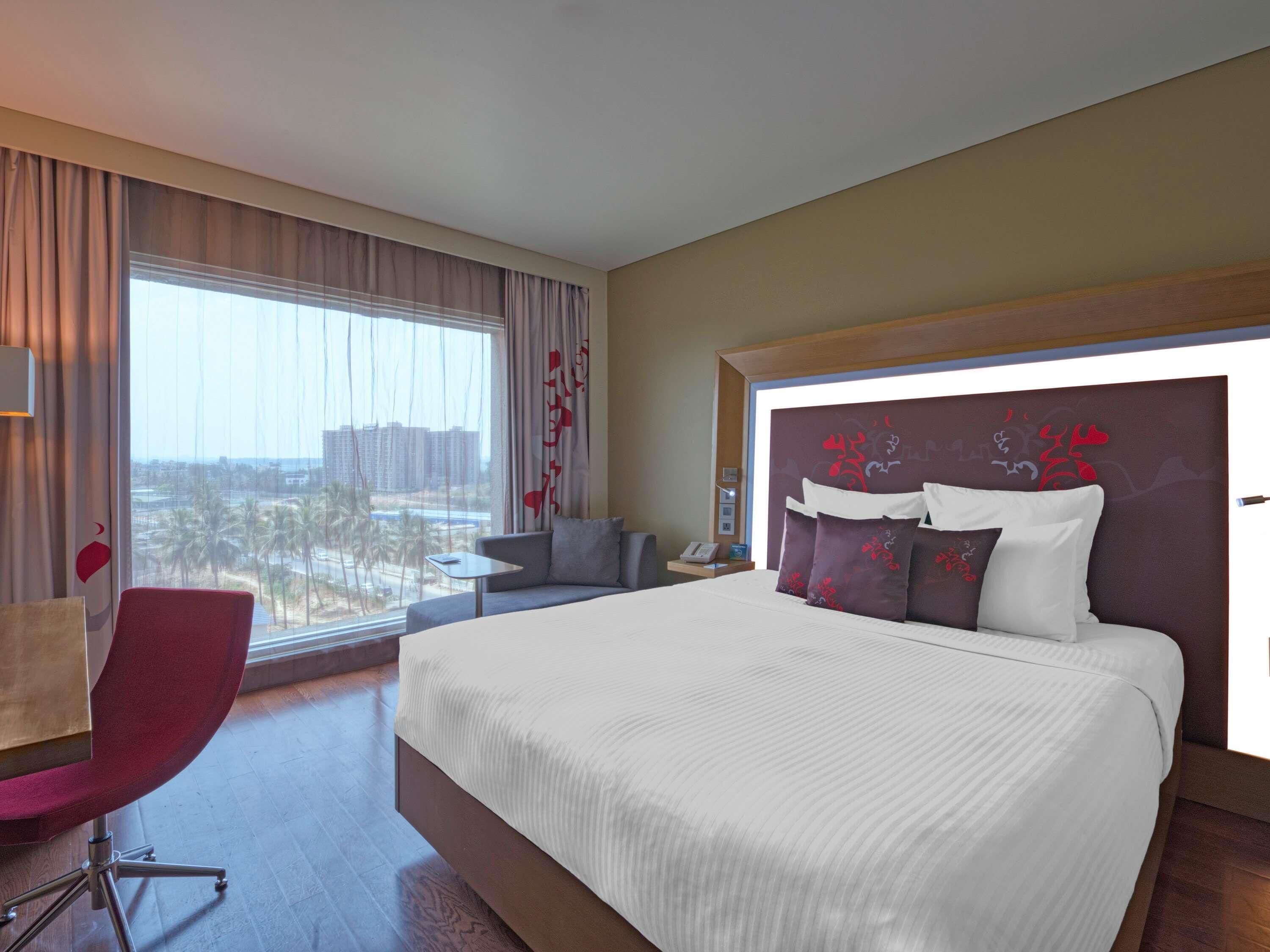 Ein Hotelzimmer mit einem großen Bett, einem Schreibtisch, einem Stuhl und Blick auf Palmen durch das Fenster.