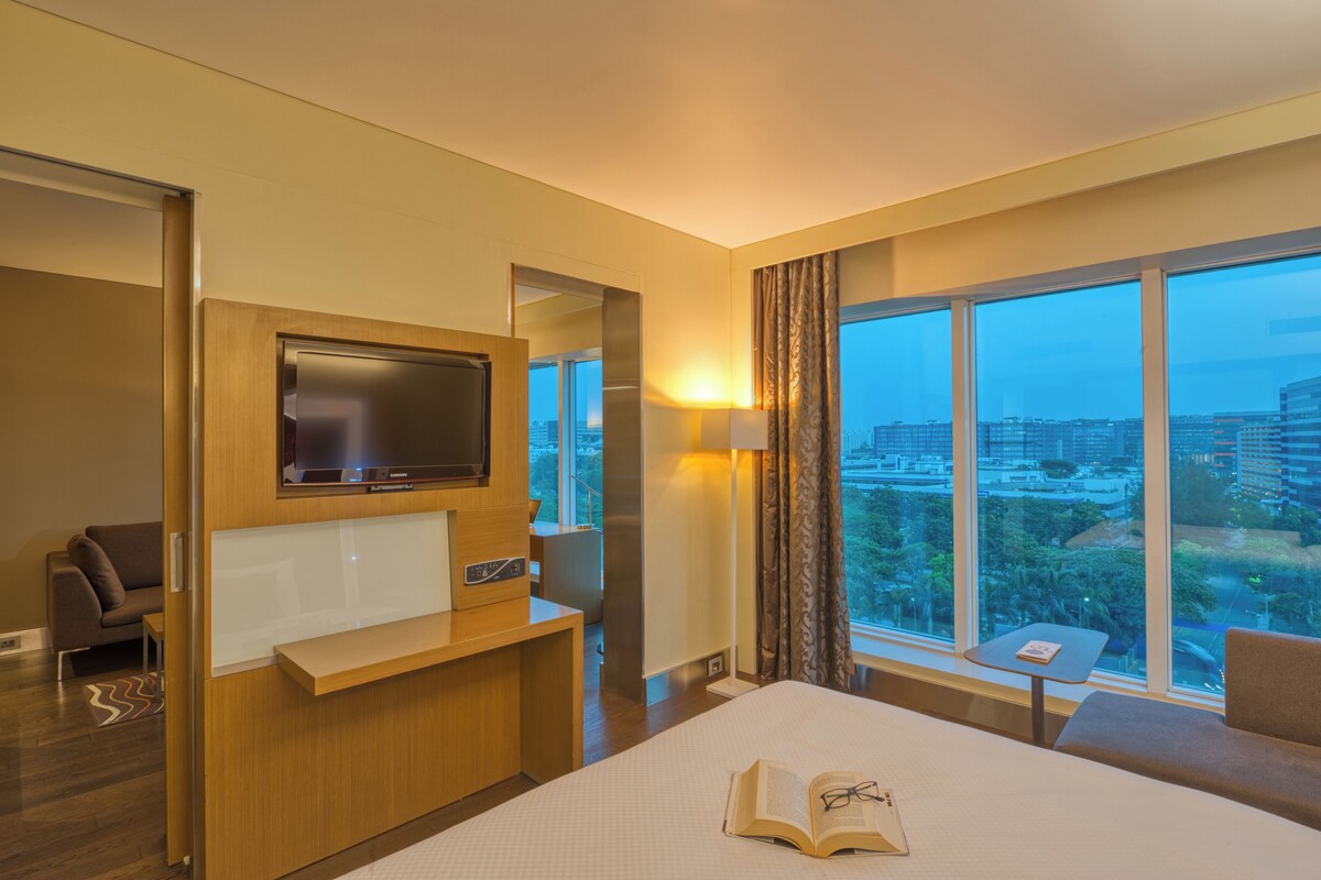 A hotel room with a bed, a TV, a desk, a chair, a small table, and a large window with a city view.