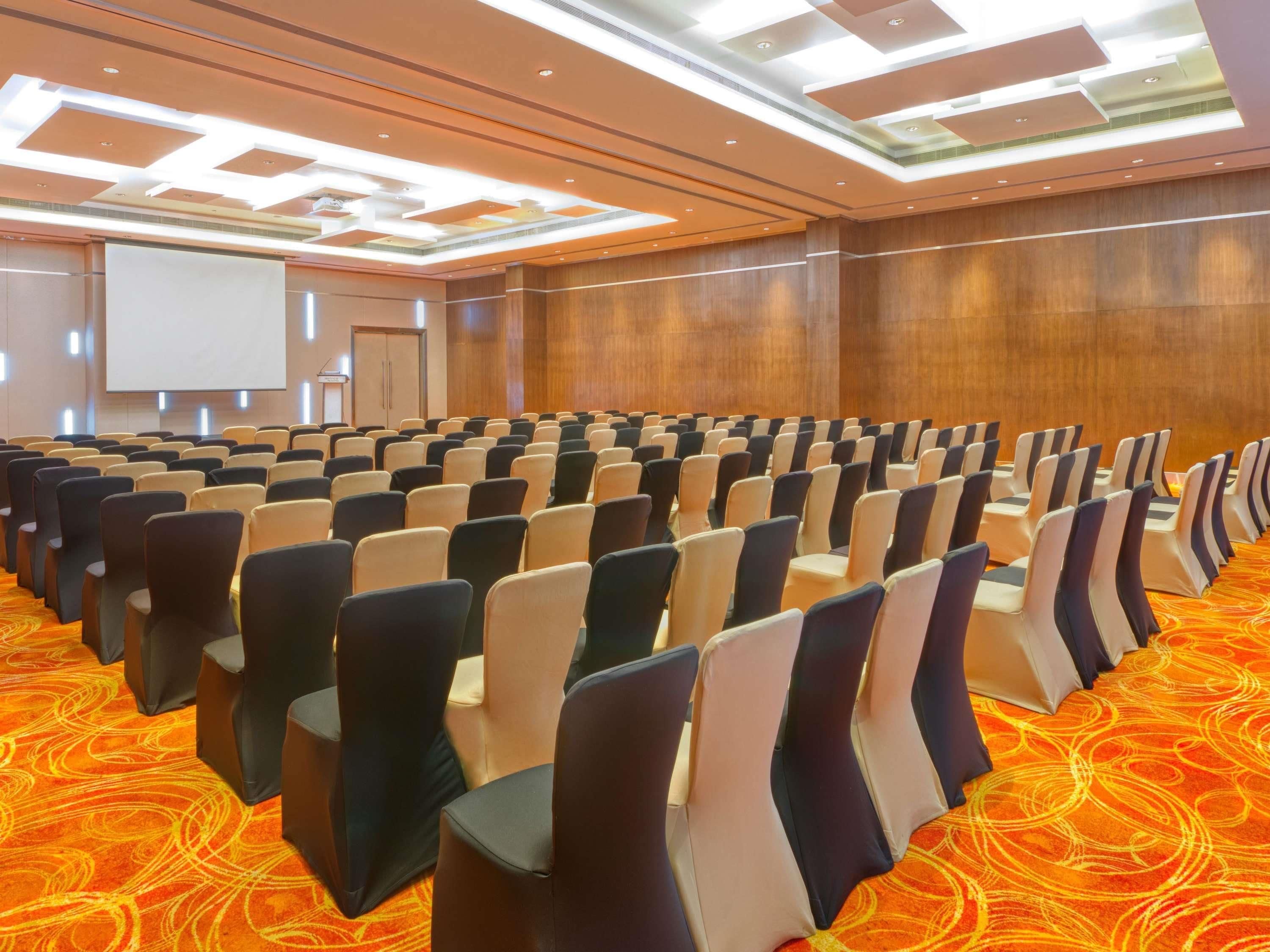 Un salón de conferencias con hileras de sillas, una pantalla de proyector y una pared de madera.