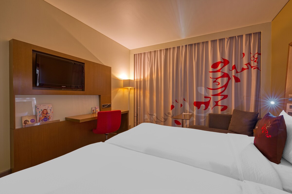 A hotel room with a large bed, a TV mounted on the wall, a desk with a chair, and a window with curtains.
