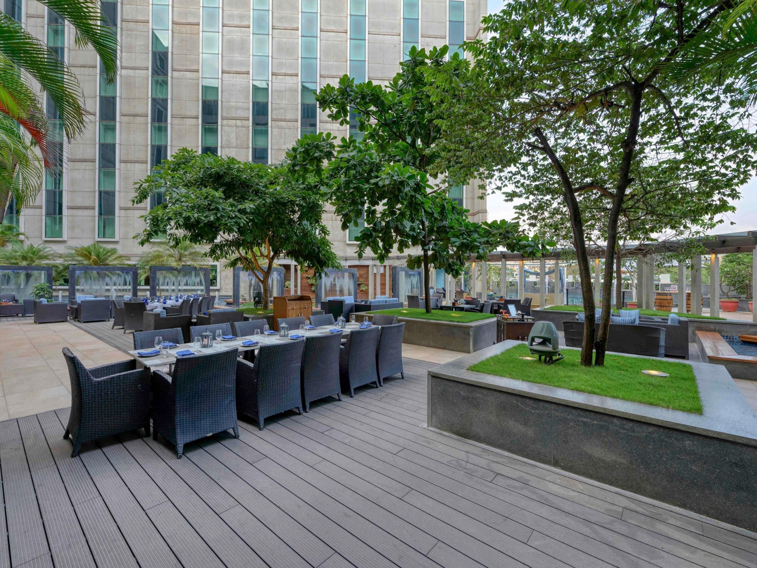 Un espace extérieur aménagé pour les repas, avec des tables et des chaises, entouré de verdure et avec un bâtiment en arrière-plan.