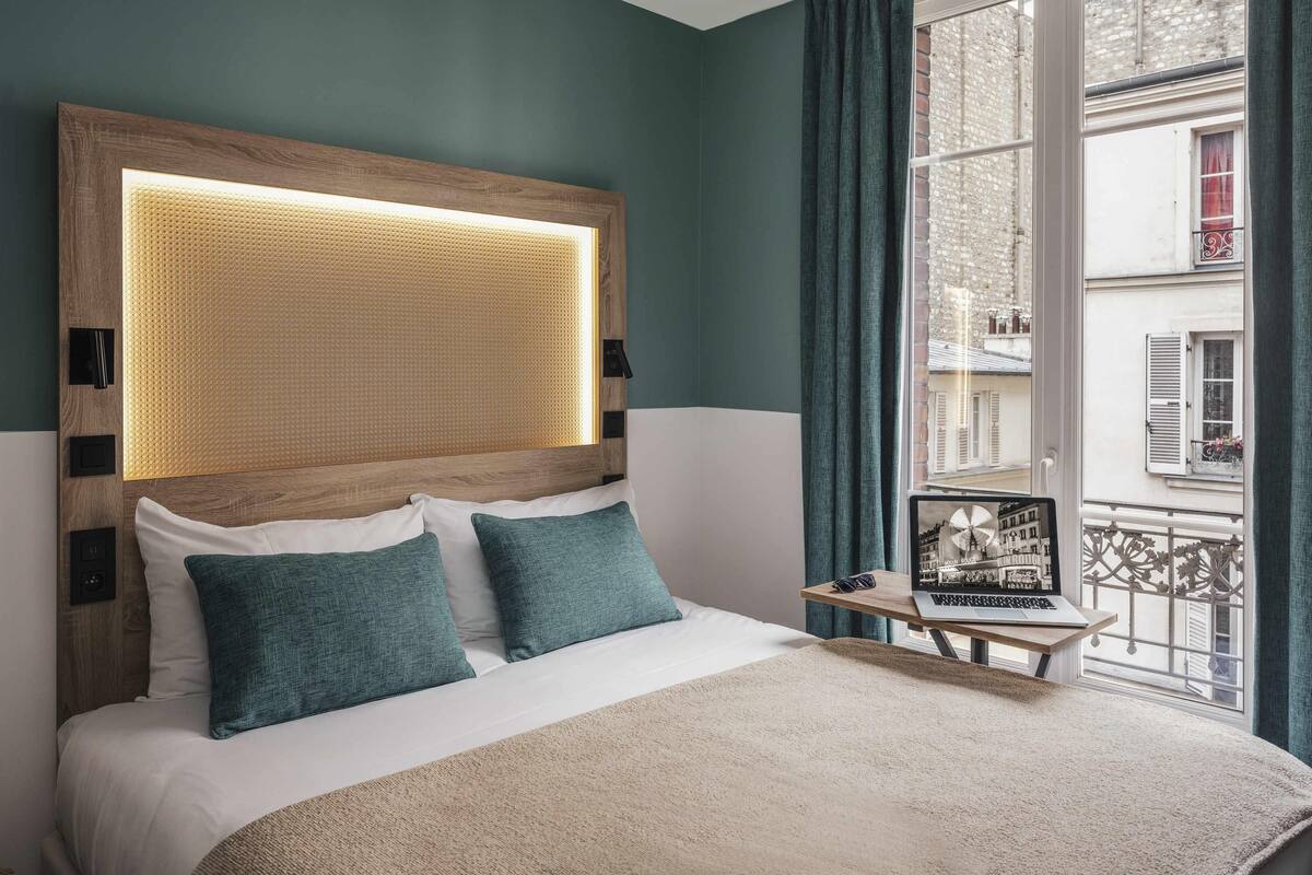 Un lit bien fait, avec une literie blanche et turquoise, une tête de lit en bois ornée d’un panneau tissé, et une petite table sur laquelle se trouvent un ordinateur portable et un cadre photo.
