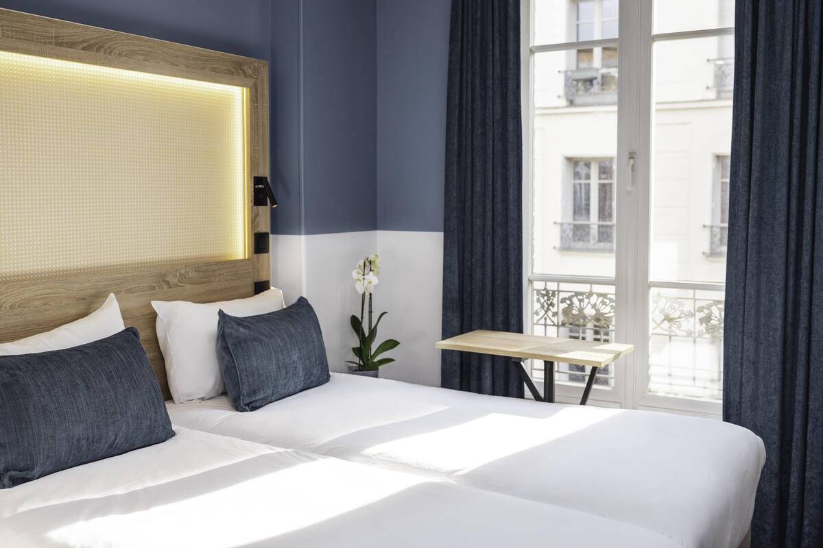 Une chambre d’hôtel avec un grand lit, une petite table, une orchidée en pot et une vue sur les immeubles par la fenêtre.